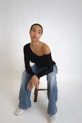 Vaenn Remsen One Shoulder Sheer Knit in black, worn by model seated on a stool with blue jeans