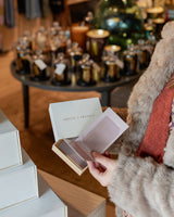 A woman in a fur coat opening an Amelie & Frank's gift card box in the boutique, the blush card lifted from cream board, Trudon candles and a Christmas tree behind her