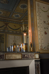 Trudon Cabinet des Miroirs candles in silver mirrored glass on a marble mantelpiece beneath a painted ceiling, beside a lit taper