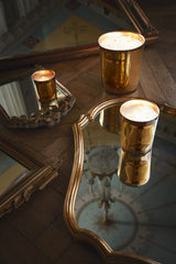 Trudon Galerie des Flammes candle in gold mirrored glass, lit on a gilt-framed mirror on a parquet floor