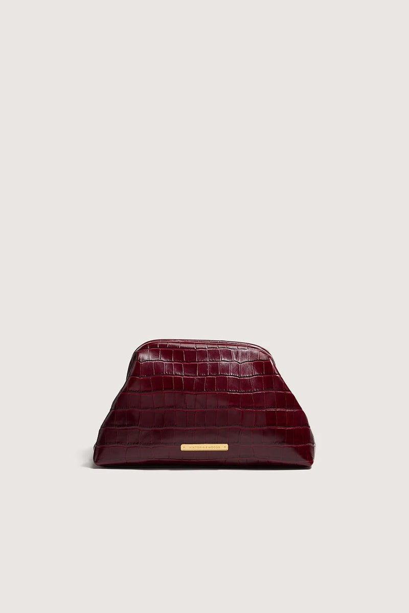 Wine-red croc-embossed leather clutch with a gold bar plaque, front view on a pale grey background